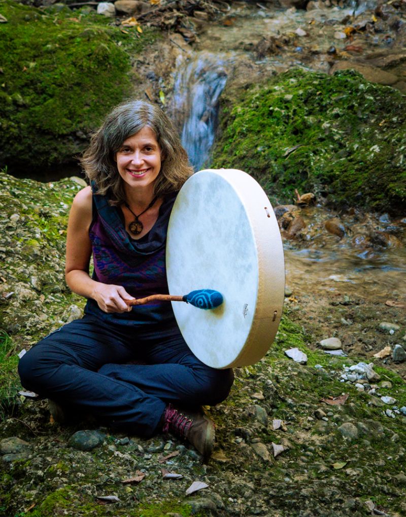Sandra Meisenburg, hier auf Moos sitzend, in der Hand eine schamanische Trommel, im Hintergrund kleiner Wasserfall am Berghang